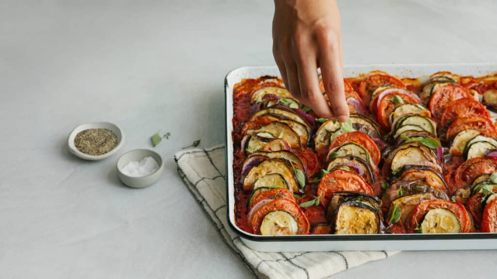 Hand sprinkling fresh herbs over a baked ratatouille dish featuring layers of eggplant, zucchini, and tomatoes, served in a white baking tray, with bowls of spices and salt in the background.