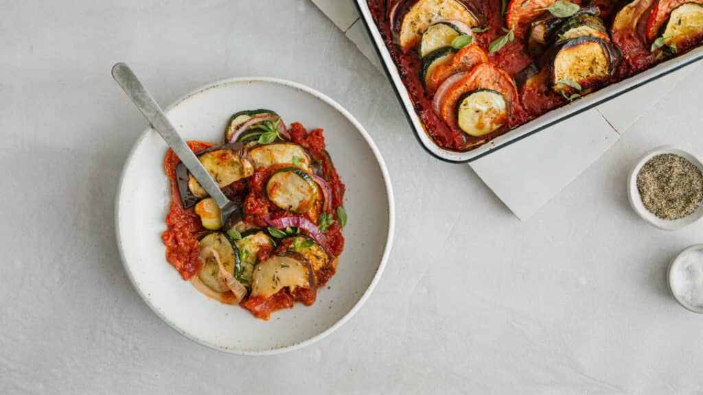 Colorful vegetable ratatouille served in a bowl with a fork, alongside a baking dish of the same dish. The ratatouille features layers of eggplant, zucchini, and bell peppers in a rich tomato sauce, garnished with fresh herbs.