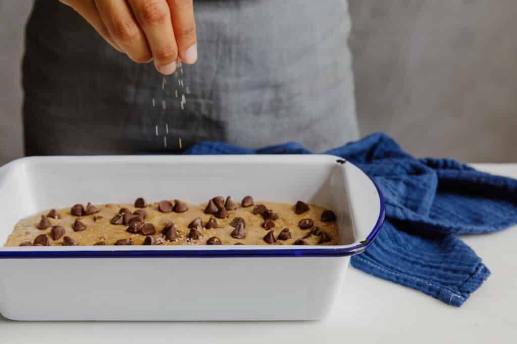 Hand sprinkling salt over a batter topped with chocolate chips in a white baking dish, with a blue kitchen towel in the background.