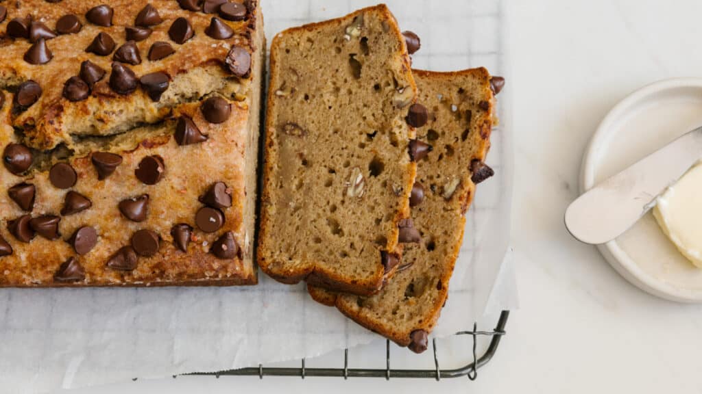 Sliced chocolate chip banana bread on a wire rack, showcasing a golden brown crust and chocolate chips on top, accompanied by a plate of butter and a butter knife.