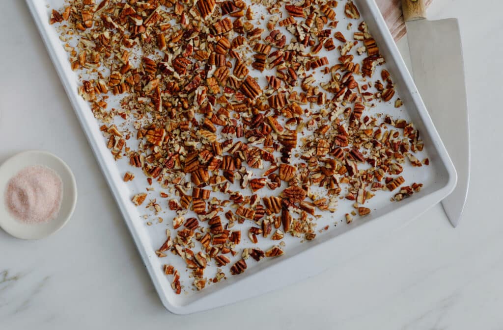 Chopped and toasted pecans spread on a baking sheet, accompanied by a small dish of pink Himalayan salt and a kitchen knife, ideal for enhancing recipes and adding flavor to dishes.