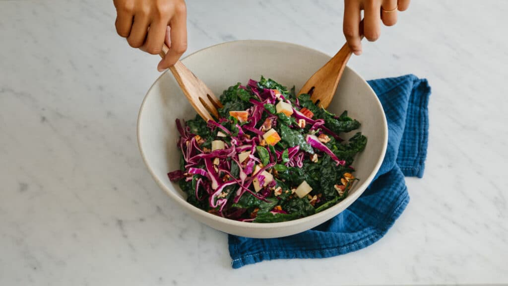 Fresh kale and red cabbage salad being tossed with wooden serving utensils in a large bowl, placed on a marble countertop with a blue cloth underneath.