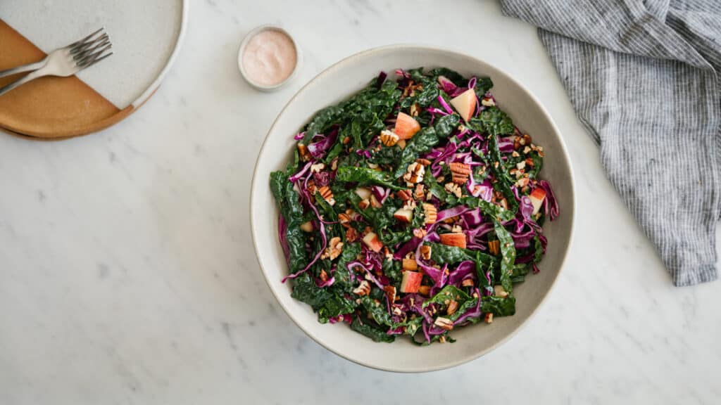 Colorful kale salad with red cabbage, diced apples, and chopped pecans in a large bowl, served on a marble countertop alongside a rustic plate and a small bowl of salt. Ideal for healthy eating and nutritious meal options.