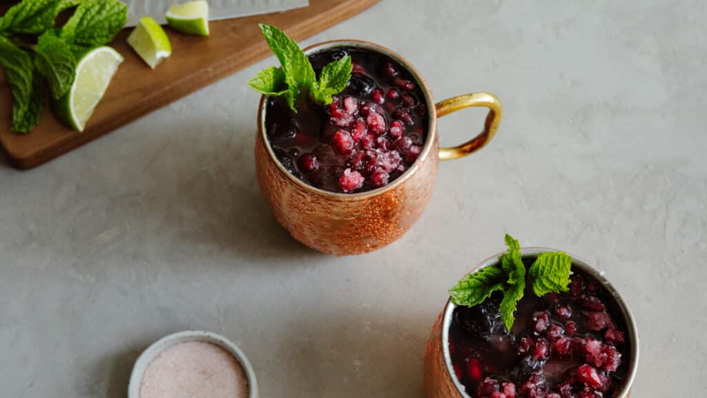 Copper mugs filled with a berry cocktail, garnished with fresh mint leaves, alongside lime wedges and a small bowl of pink salt on a textured surface.