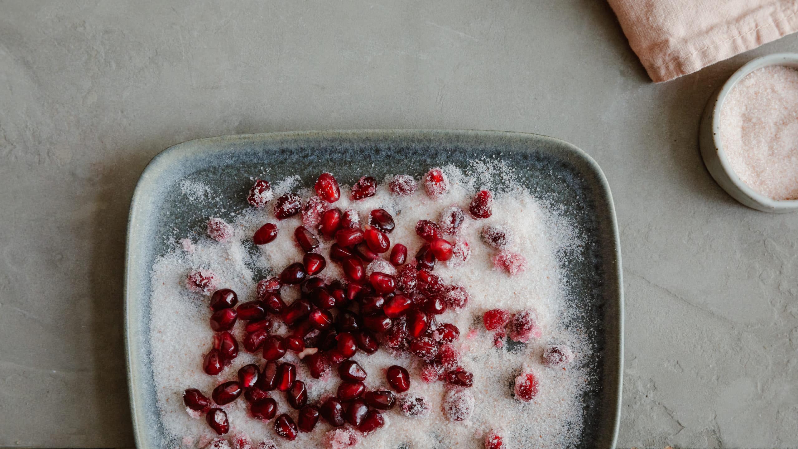 Pomegranate seeds sprinkled with coarse pink salt on a blue plate, with additional salt in a small bowl nearby, showcasing a vibrant and visually appealing culinary presentation.