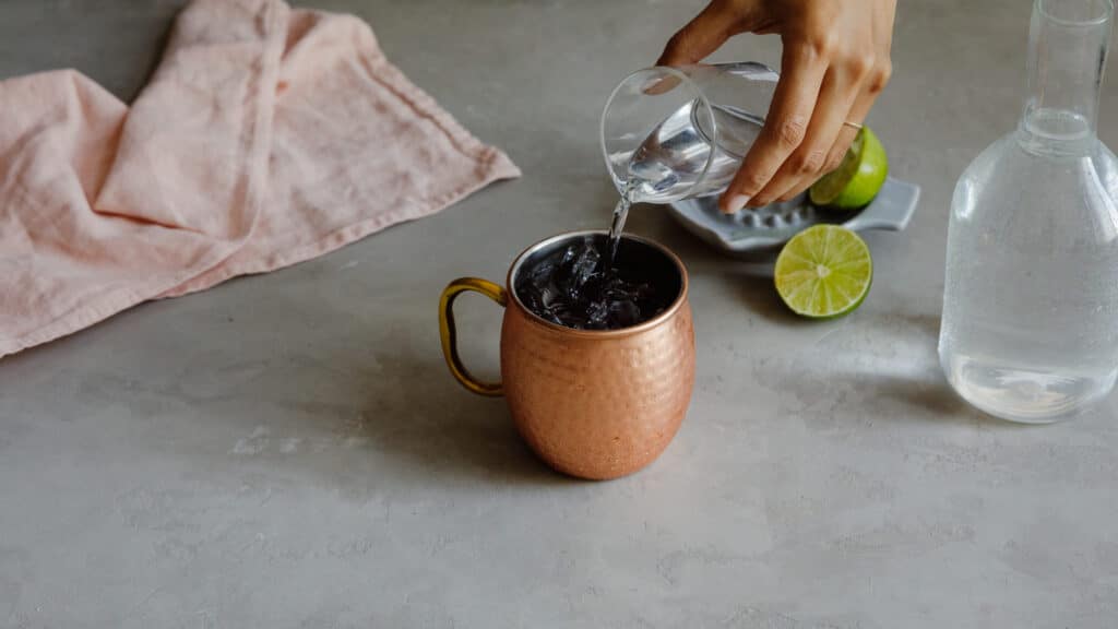 Hand pouring water into a copper mug filled with ice, surrounded by a pink cloth, a plate with lime halves, and a glass bottle of water on a gray countertop.