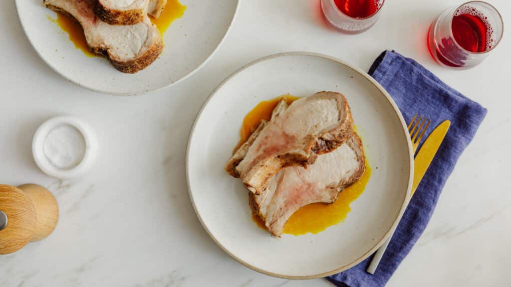 Plated slices of succulent roasted pork with a savory sauce, accompanied by a purple linen napkin and garnished with a wooden salt cellar and two glasses of red beverage.