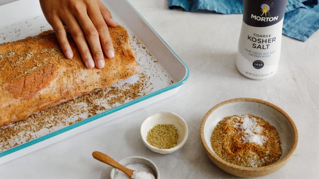 Hand applying a spice rub to a seasoned pork loin on a baking tray, with containers of coarse kosher salt and various spices in the foreground.