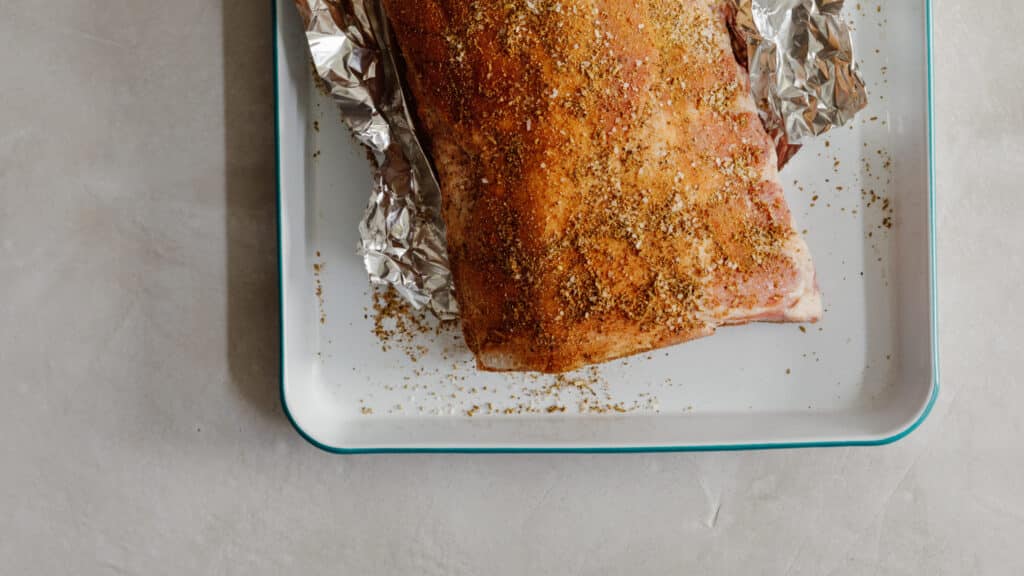 Raw pork belly seasoned with spices, resting on a white tray lined with aluminum foil, ready for cooking.