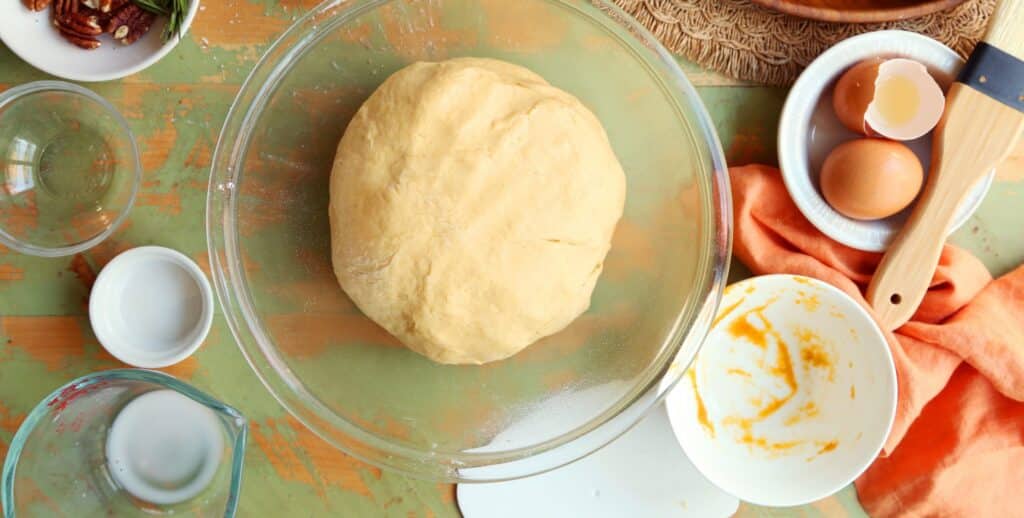 Homemade dough resting in a glass bowl, surrounded by ingredients including eggs, pecans, and measuring cups on a rustic kitchen table.