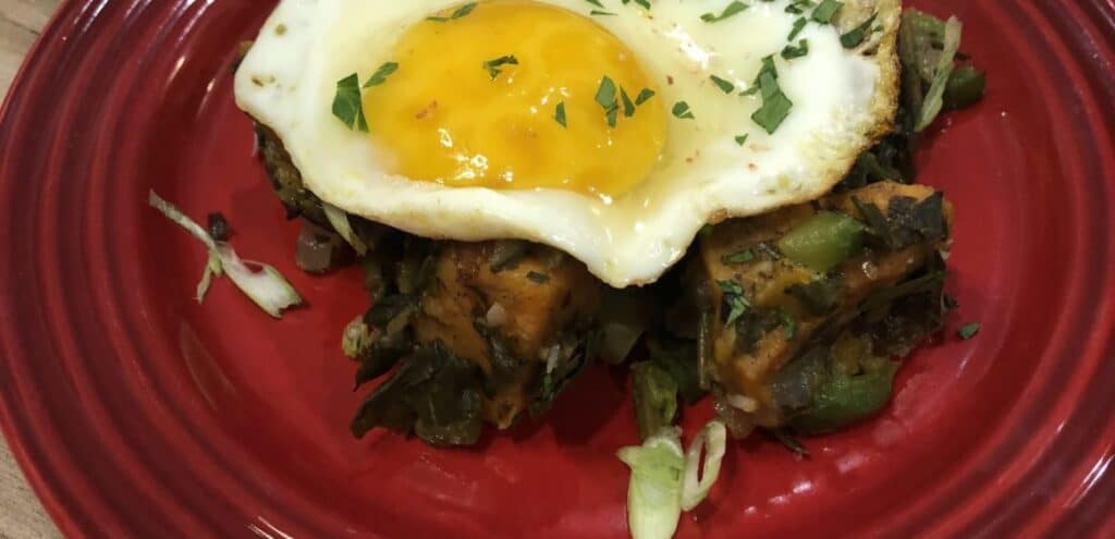 A vibrant dish featuring a sunny-side-up egg on top of seasoned sautéed greens and diced sweet potatoes, garnished with fresh herbs, served on a red plate.