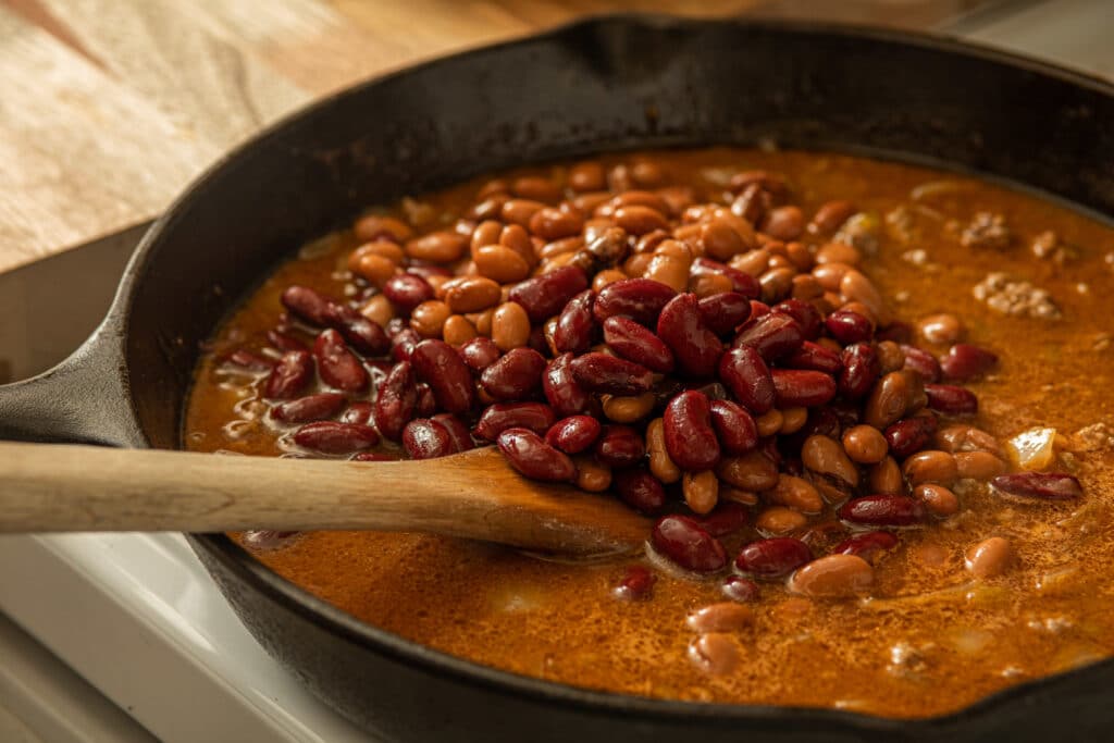A cast iron skillet filled with a hearty bean mixture, featuring red kidney beans and pinto beans simmering in a savory sauce, with a wooden spoon resting in the dish.