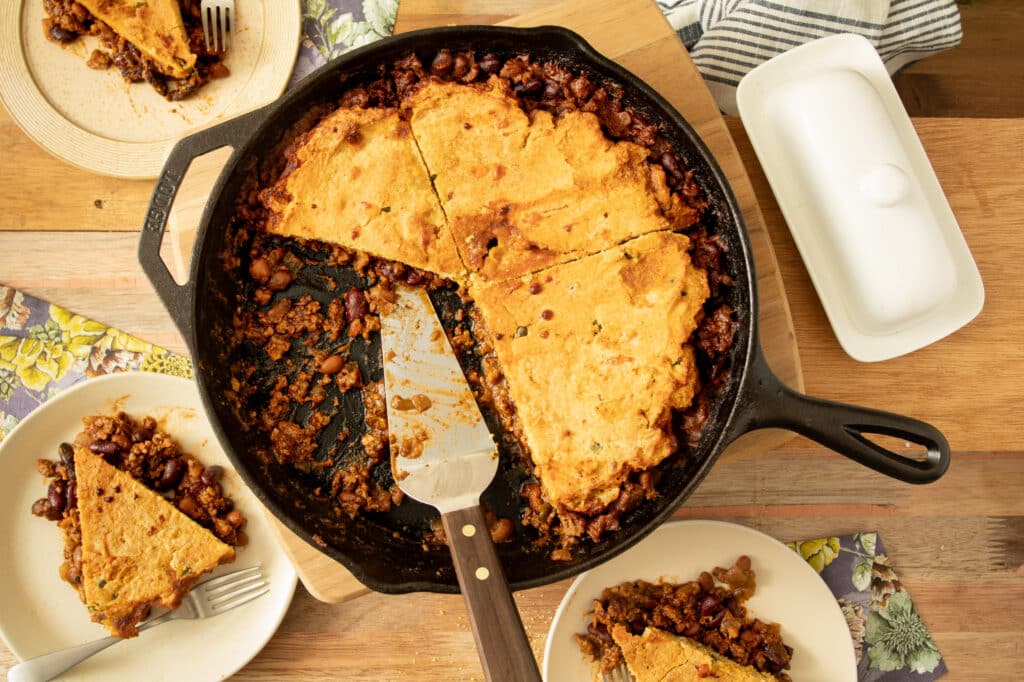 Cast iron skillet filled with cornbread topped chili, featuring slices of cornbread served on plates, showcasing a hearty and comforting meal.