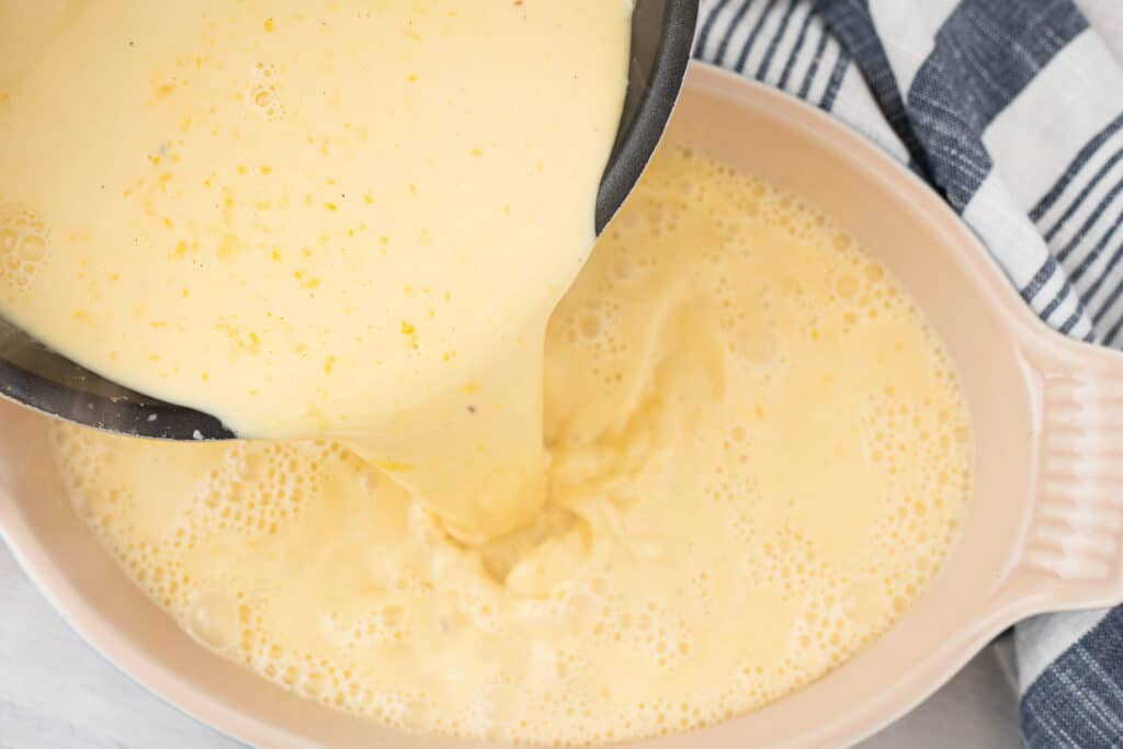 Creamy mixture being poured from a saucepan into a baking dish, showcasing a smooth, yellow batter with bubbles, ideal for recipes like custards or quiches. A striped kitchen towel is partially visible in the background.