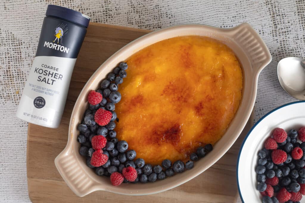 Creamy dessert topped with a caramelized layer, surrounded by fresh blueberries and raspberries, next to a container of Morton coarse kosher salt.