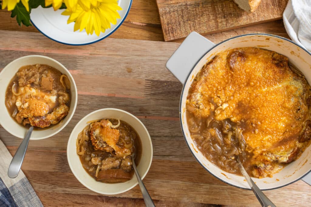 Bowl of French onion soup topped with melted cheese, served alongside a pot of the soup and slices of bread on a wooden table, with a bouquet of yellow flowers in the background.