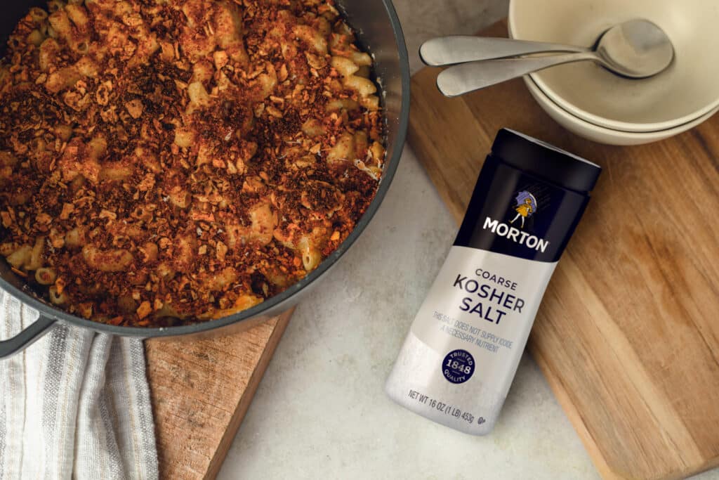 A delicious baked macaroni and cheese dish topped with crunchy breadcrumbs, beside a container of Morton Coarse Kosher Salt, set on a wooden cutting board with bowls and utensils in the background.