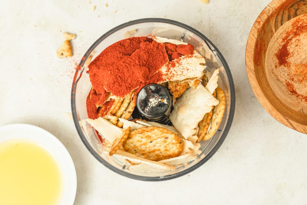 Ingredients in a food processor including crackers, red spice powder, and oil, with a wooden bowl of spices in the background. Perfect for making flavorful dips or seasonings.