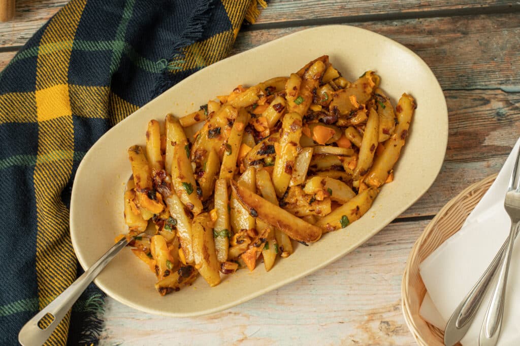 Stir-fried potato wedges seasoned with spices and garnished with chopped vegetables, served on a beige plate alongside a checkered cloth and utensils.