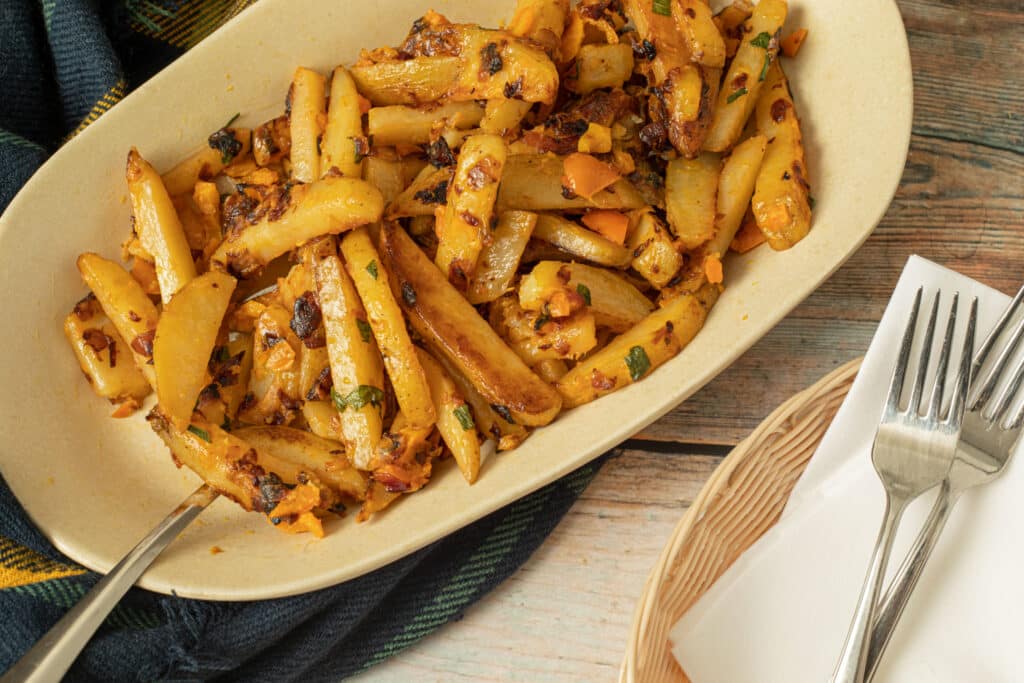 Golden-brown roasted potato wedges seasoned with herbs and spices, served in a beige dish on a rustic wooden table. A fork rests beside the dish, with a woven basket holding napkins and utensils nearby.