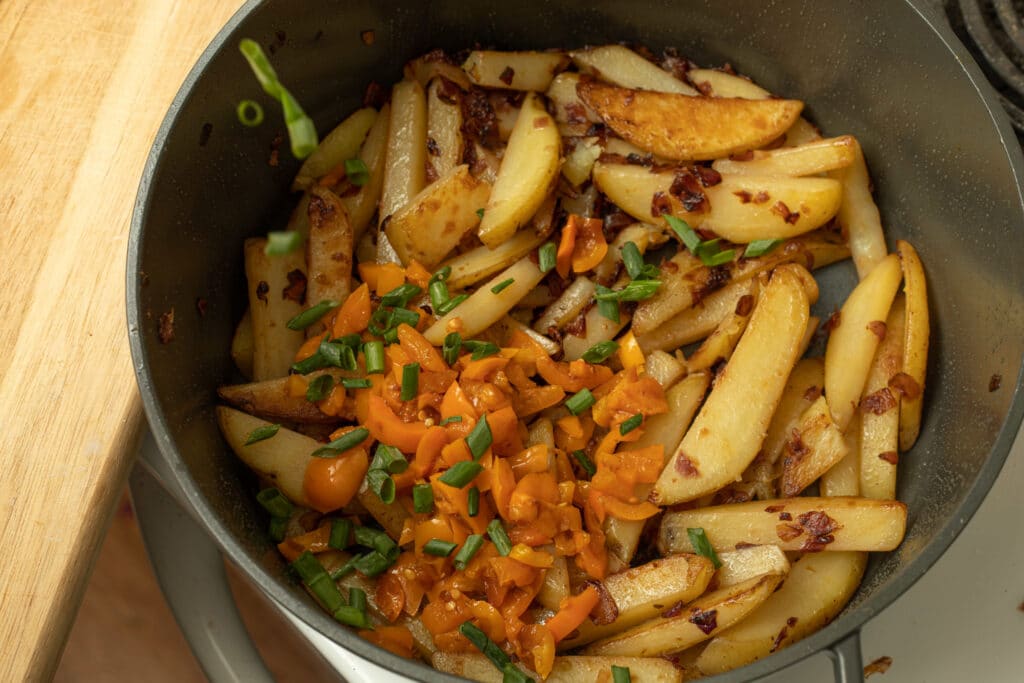 Golden-brown potato wedges seasoned and sautéed, topped with diced orange peppers and chopped green onions, served in a black pot on a wooden surface.