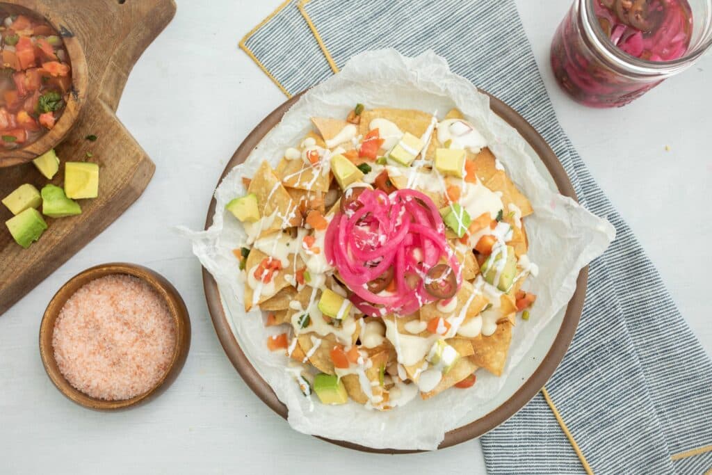 Colorful nachos topped with diced tomatoes, avocado, pickled onions, and drizzled with creamy sauce, served on a rustic plate alongside a bowl of pink Himalayan salt and fresh salsa.
