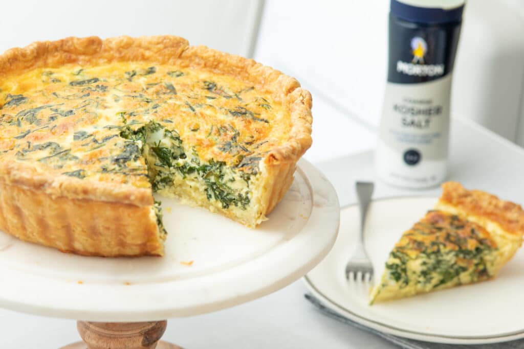 Delicious spinach quiche on a marble cake stand, featuring a golden, flaky crust and a creamy filling with fresh spinach. A slice is plated beside the quiche, garnished with a fork, and a container of kosher salt is visible in the background. Perfect for brunch or a light meal.