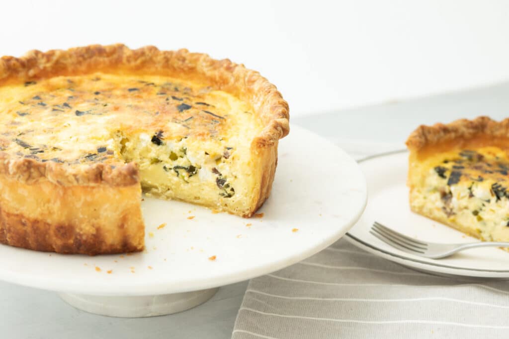 Delicious spinach and cheese quiche on a marble cake stand, showcasing a slice removed, with a fork on a plate nearby, perfect for brunch or a light meal.