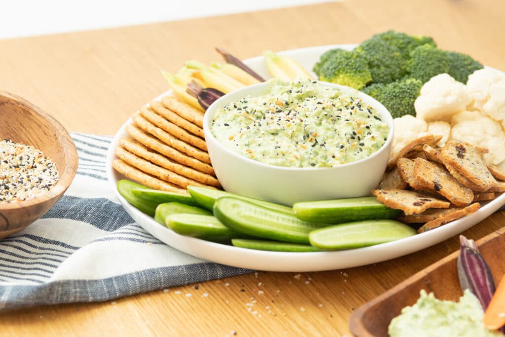 A vibrant vegetable platter featuring creamy dip in a bowl, surrounded by fresh cucumber slices, broccoli, cauliflower, and crispy crackers, with a bowl of mixed seeds in the background. Perfect for healthy snacking or entertaining guests.