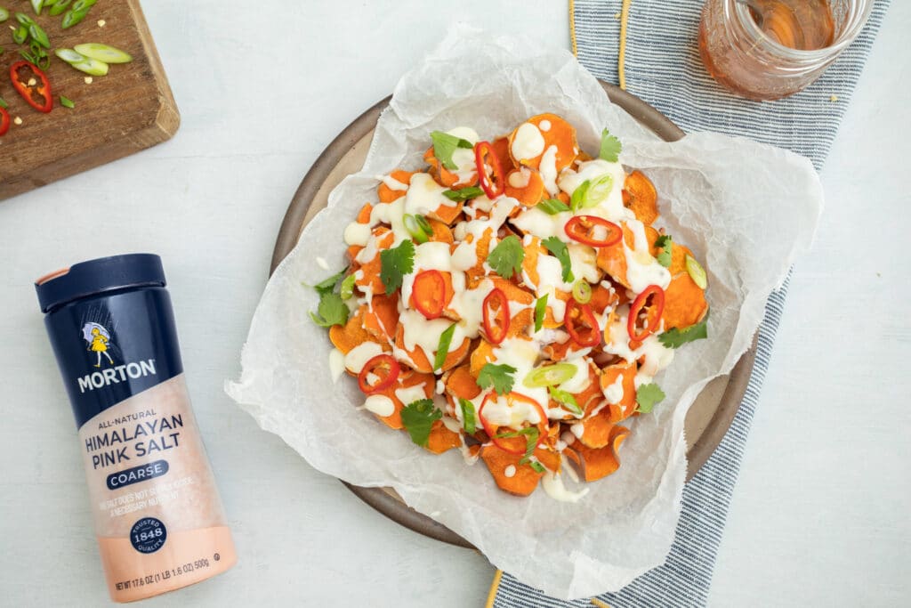 Sweet potato nachos topped with cilantro, sliced red peppers, and drizzled with creamy sauce, alongside a container of Morton Himalayan Pink Salt on a rustic table setting.