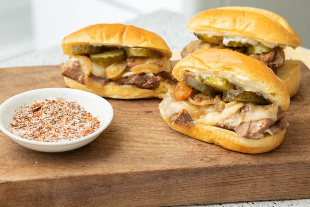Juicy beef sandwiches topped with pickles and sautéed onions, served on soft rolls, alongside a bowl of seasoned salt for added flavor.
