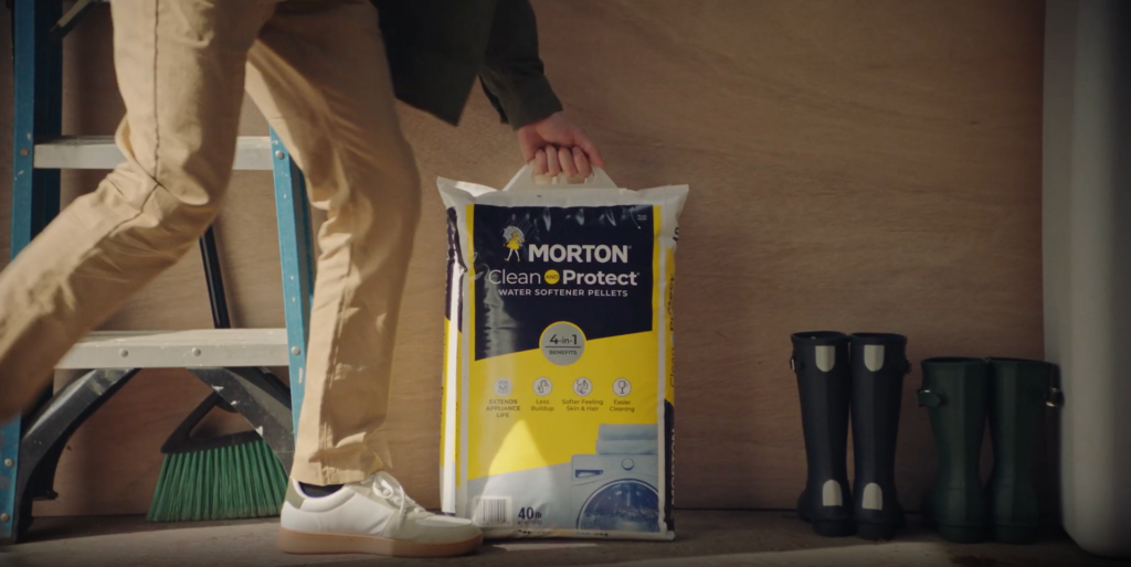 Person holding a bag of Morton Clean and Protect water softener pellets, highlighting its 4-in-1 benefits for improved appliance life, skin feel, and easier cleaning, with a broom and rubber boots in the background.