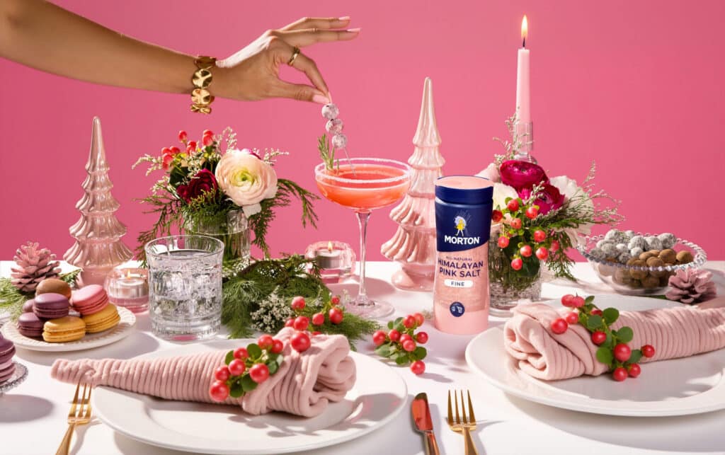 Festive dinner table setting featuring Morton Himalayan Pink Salt, a vibrant pink background, elegant floral arrangements, and a cocktail garnished with berries. The table is adorned with gold cutlery, decorative napkins, and an assortment of colorful macarons, creating a cheerful holiday ambiance.