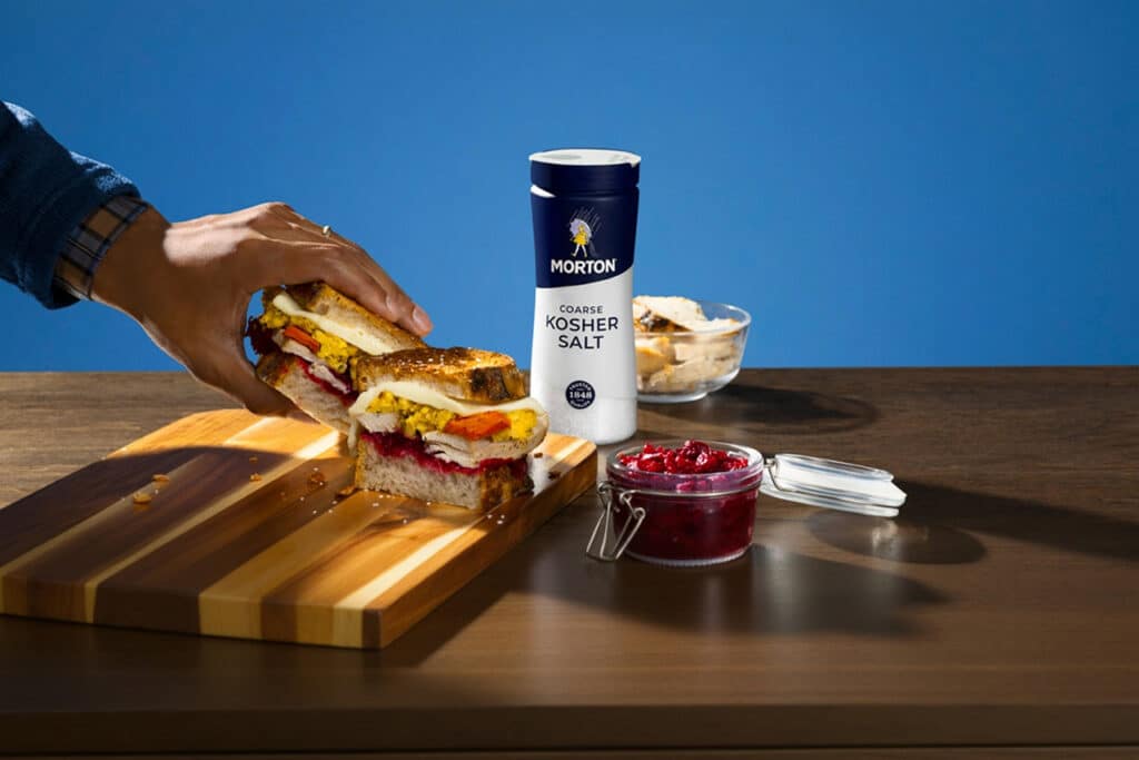 Hand reaching for a hearty sandwich stacked with turkey, eggs, and vegetables, placed on a wooden cutting board next to Morton Coarse Kosher Salt and a jar of cranberry sauce, with a blue background.
