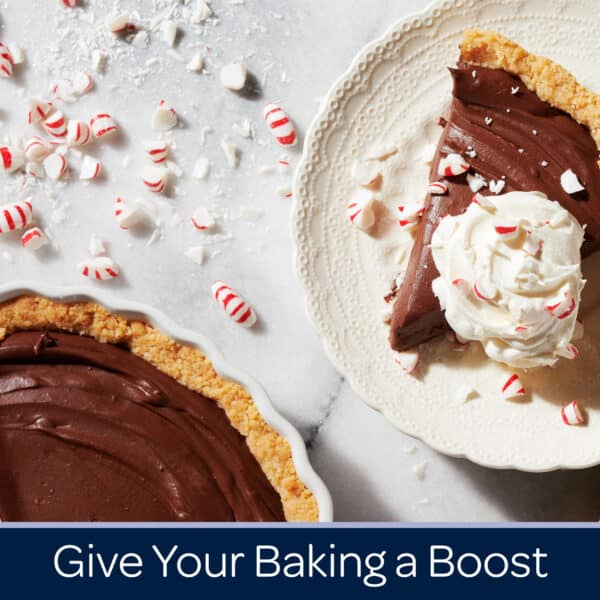 Chocolate cream pie slice topped with whipped cream and crushed peppermint, surrounded by festive peppermint candies on a marble surface, with a whole pie in the background. Perfect for holiday baking inspiration.