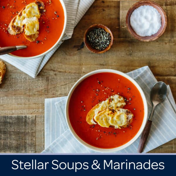 Bowl of vibrant tomato soup topped with toasted bread and sprinkled with black pepper, accompanied by a small bowl of herbs and a pinch of salt, set on a wooden table.