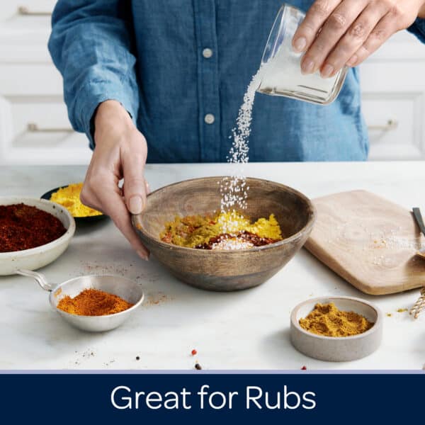 Person preparing a spice blend for rubs by pouring salt into a wooden bowl filled with various spices, surrounded by small bowls of additional spices on a marble countertop.