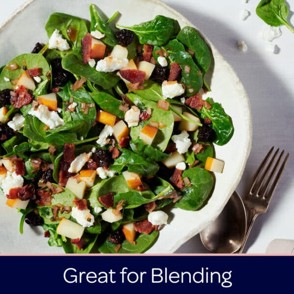 Fresh spinach salad topped with diced apples, crumbled goat cheese, crispy bacon, and dried cherries in a textured bowl, garnished with chopped onions, perfect for a nutritious meal.