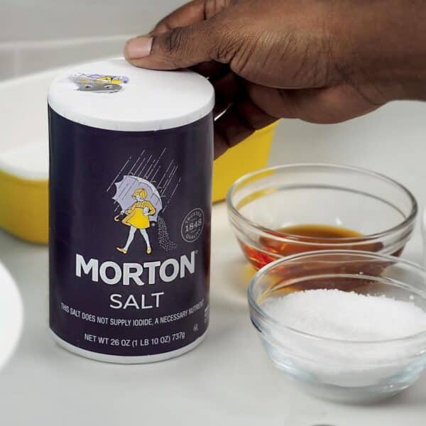 Morton Salt container held in a hand, surrounded by bowls of various ingredients, including brown liquid and white granulated salt, on a kitchen countertop.
