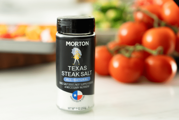 Morton Texas Steak Salt bottle in focus, featuring an all-natural label, with fresh tomatoes blurred in the background. Ideal for enhancing the flavor of steak and other meats, highlighting its Texas origin.