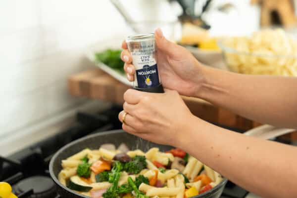 Person adding sea salt to a pan of mixed vegetables and pasta while cooking in a modern kitchen. The image highlights the process of seasoning a healthy meal, emphasizing fresh ingredients and culinary techniques.