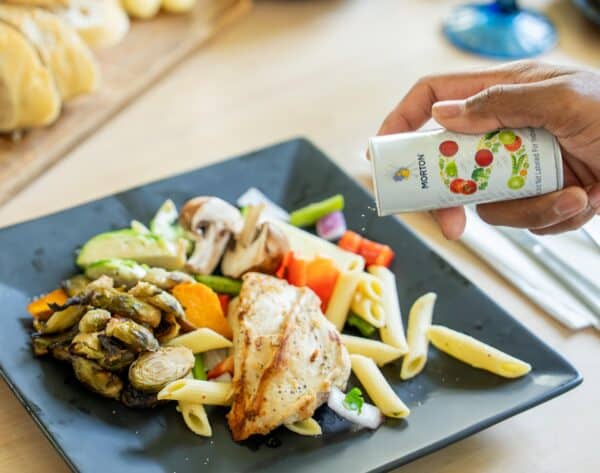 A hand sprinkles seasoning from a Morton's container over a plate of grilled chicken, pasta, and colorful vegetables including zucchini, bell peppers, and mushrooms, set on a dark square plate with a wooden table in the background.