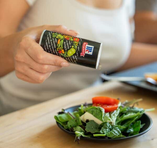 Woman seasoning a fresh salad with McCormick spices, showcasing vibrant greens and tomatoes on a black plate.