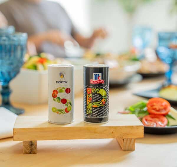 Morton and McCormick seasoning containers displayed on a wooden stand, with a colorful salad and fresh vegetables in the background, highlighting a vibrant dining setting.