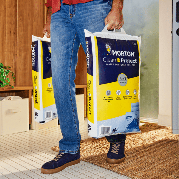 Person carrying two 40-pound bags of Morton Clean and Protect water softener pellets, showcasing a 4-in-1 benefits label. The setting features a modern home interior with a tiled floor, emphasizing the product's role in extending appliance life and reducing buildup.