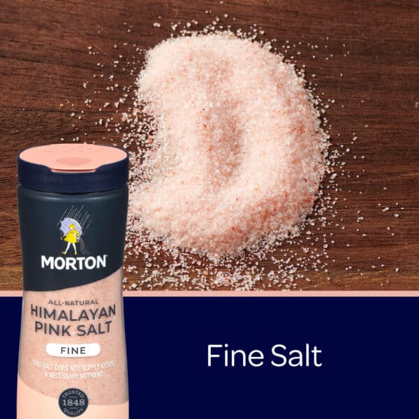 Morton Himalayan Pink Salt in a container with fine salt granules scattered on a wooden surface, highlighting its natural pink color and texture.