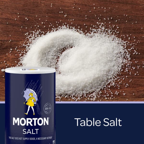 Container of Morton table salt on a wooden surface, with granules of salt arranged in a heart shape around it. Ideal for seasoning and enhancing flavor in cooking and baking.