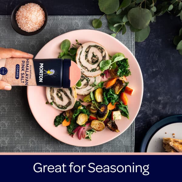 A hand holding a container of Morton Himalayan pink salt, sprinkling it over a colorful plate of sliced rolled chicken with spinach, roasted vegetables, and greens, showcasing a vibrant and healthy meal.