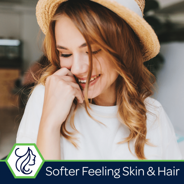 Smiling woman with wavy hair wearing a straw hat, showcasing soft skin and hair in a bright indoor setting. The image emphasizes beauty and self-care, promoting products for enhanced skin and hair softness.