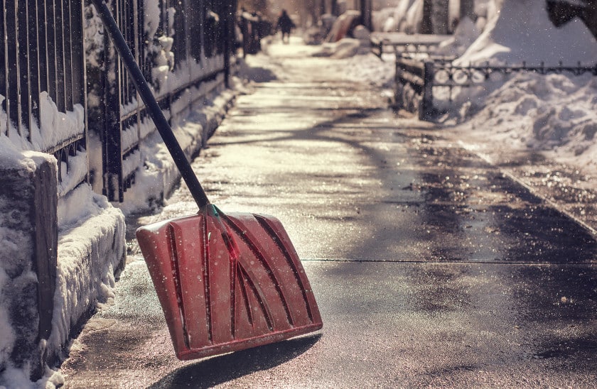 A red snow shovel resting against a cleared urban sidewalk after a snowstorm, with snow piled along iron fencing and a sunlit path stretching into the background.